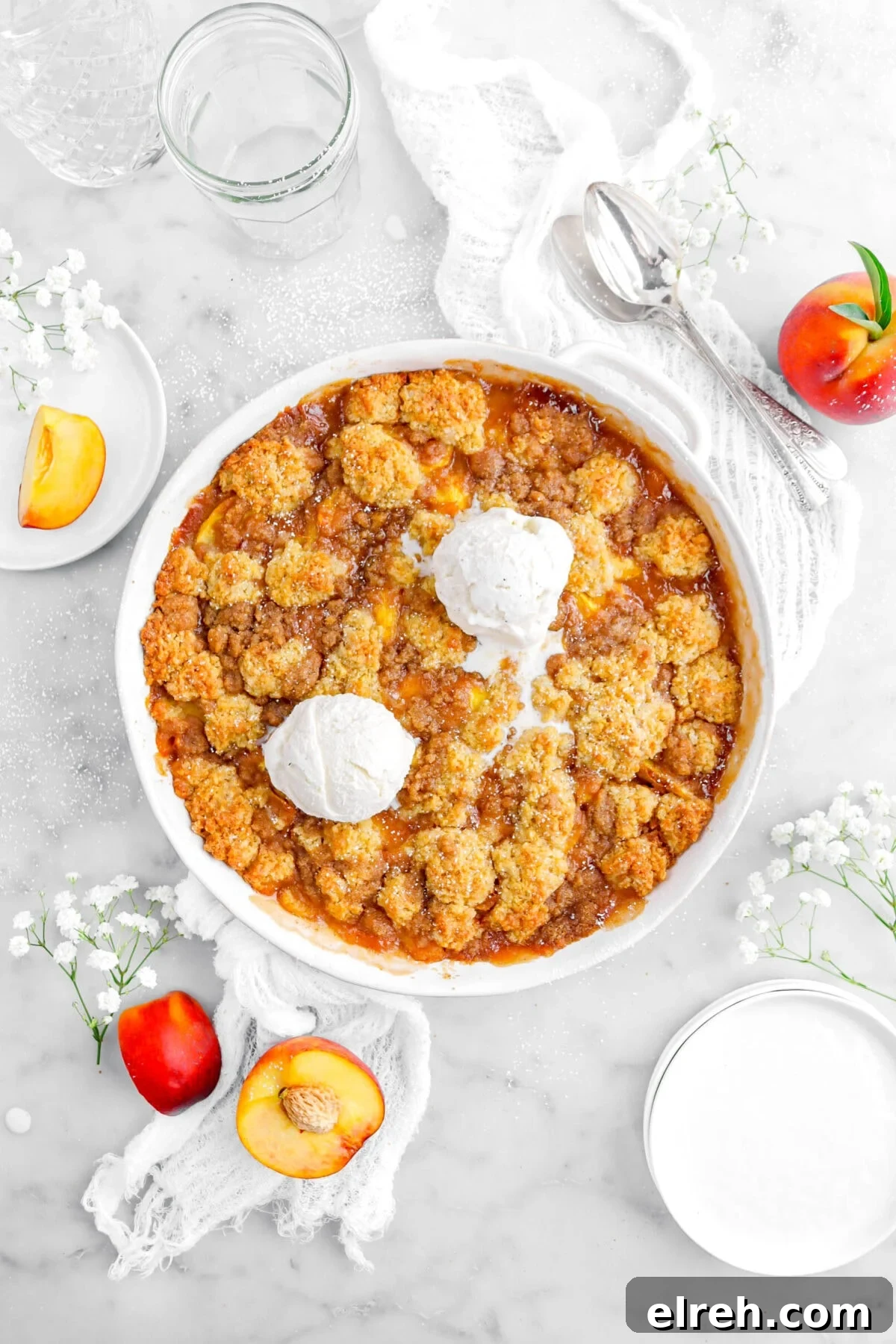 Peach Cobbler Crisp with Brown Sugar and Cinnamon 6 peach cobbler in round casserole with two scoops of vanilla ice cream top with peaches, white flowers, plates, and two spoons around on marble surface.