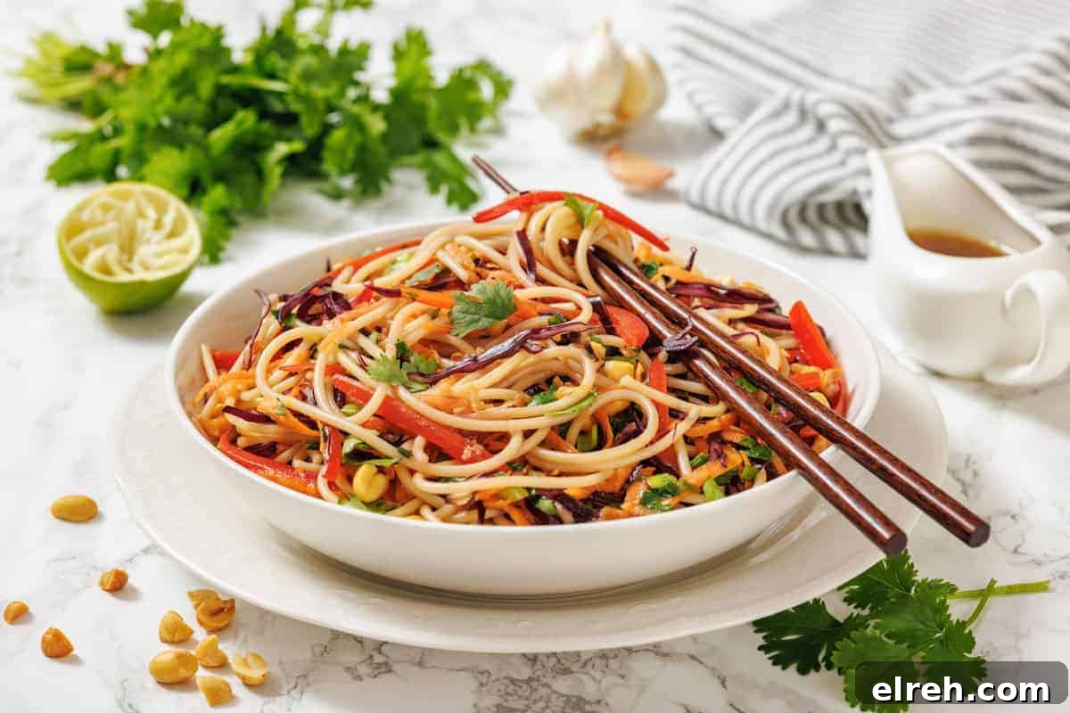 A bowl of Asian-inspired noodle salad with vegetables, garnished with cilantro, chopsticks on top, and ingredients like lime, peanuts, and fresh herbs nearby.