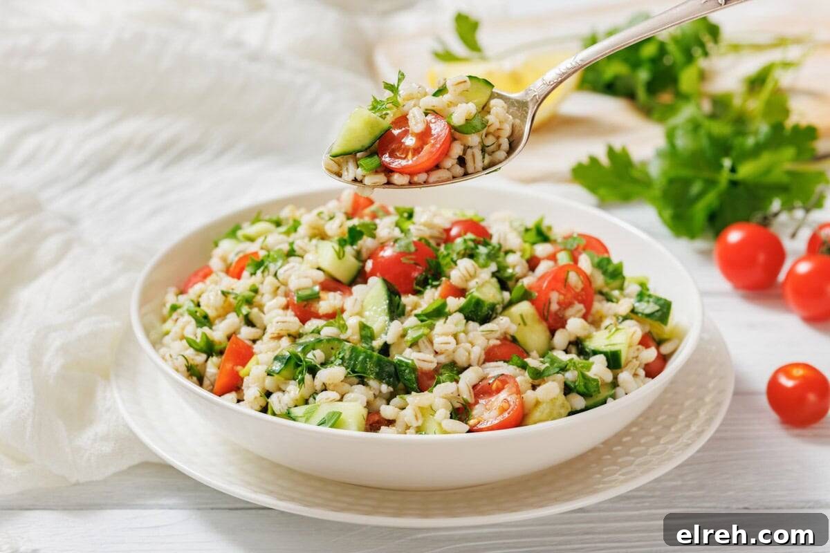 A bowl of barley salad with cherry tomatoes, cucumber, and parsley sits on a plate, with a spoonful of salad being held above the bowl.