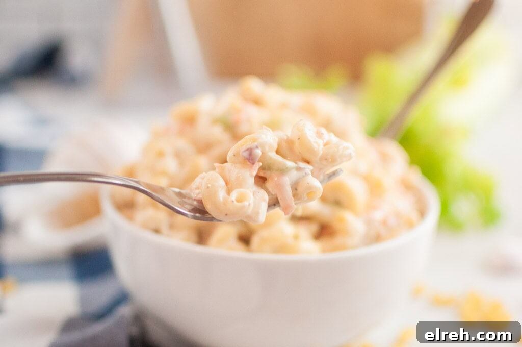 A close-up shot of a fork lifting a portion of macaroni salad, with a bowl of the salad in the blurred background.