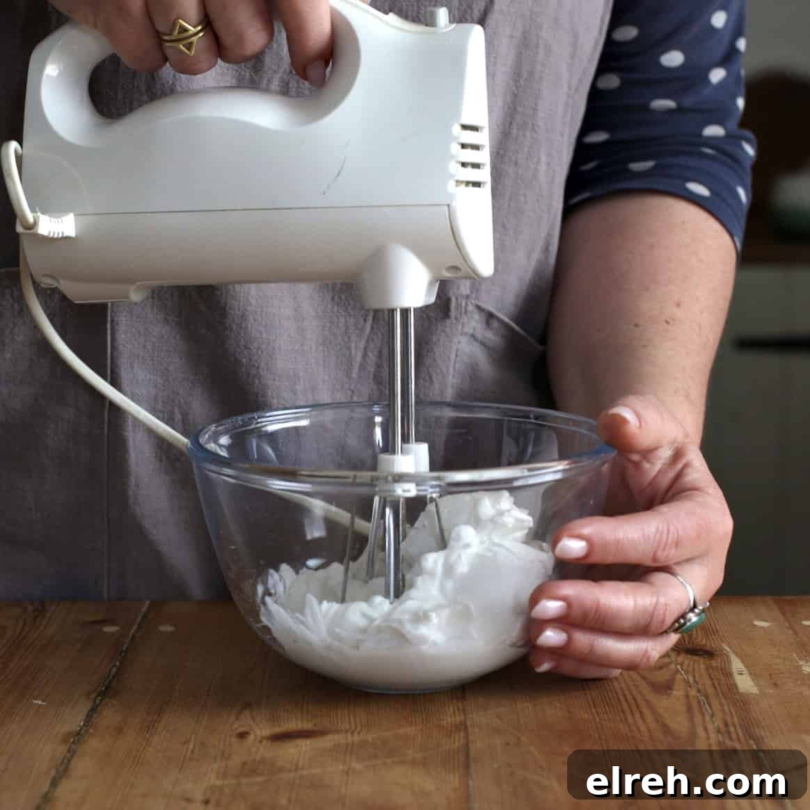 Woman in grey using a white electric whisk to beat canned coconut milk in a glass mixing bowl