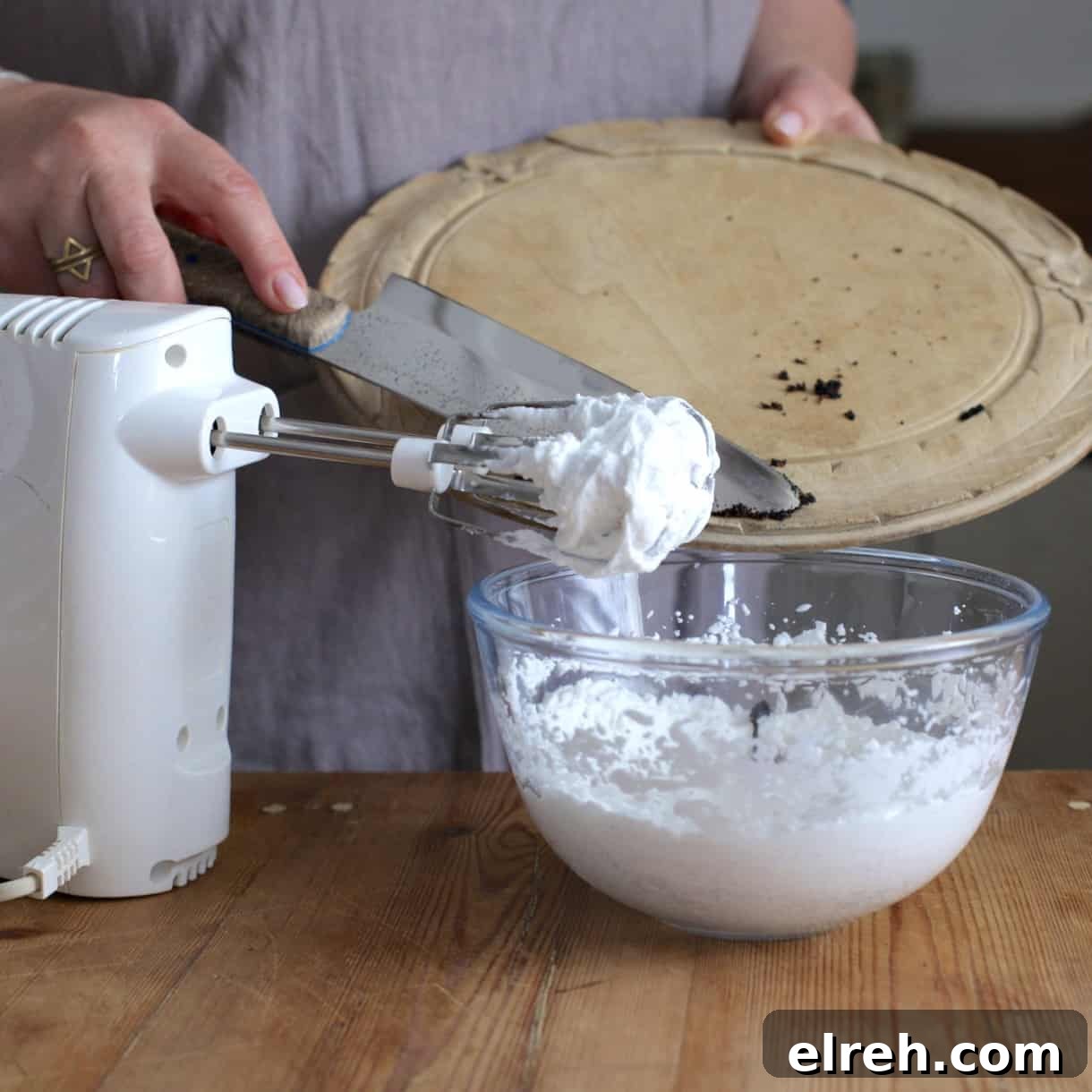 Hand mixer and glass bowl of whipped coconut cream with a woman scraping vanilla beans from a wooden board into the bowl of whipped cream