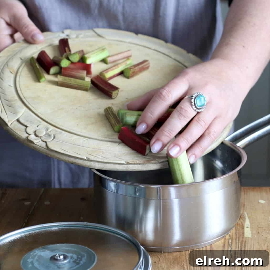 Woman sliding cut pieces of raw rhubarb from a wooden chopping board into a silver saucepan