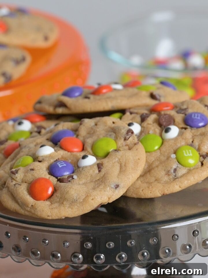 Baked Halloween chocolate chip cookies decorated with colorful M&M's and candy eyeballs on a black cake stand.
